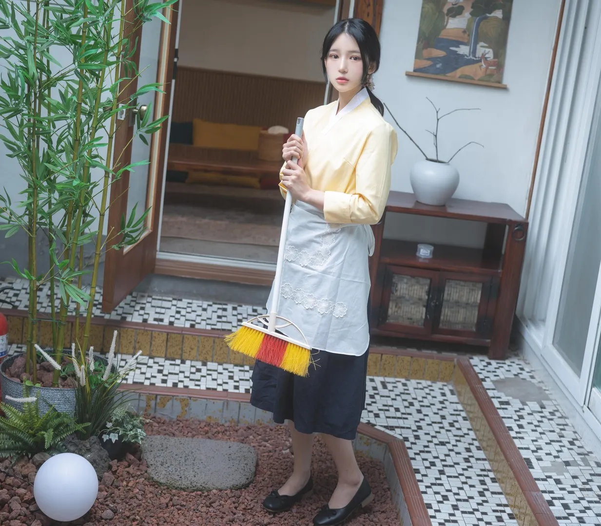 Yeha(예하)穿浅黄色韩服上衣和深色裙装,手持扫帚站在室内庭院场景中
