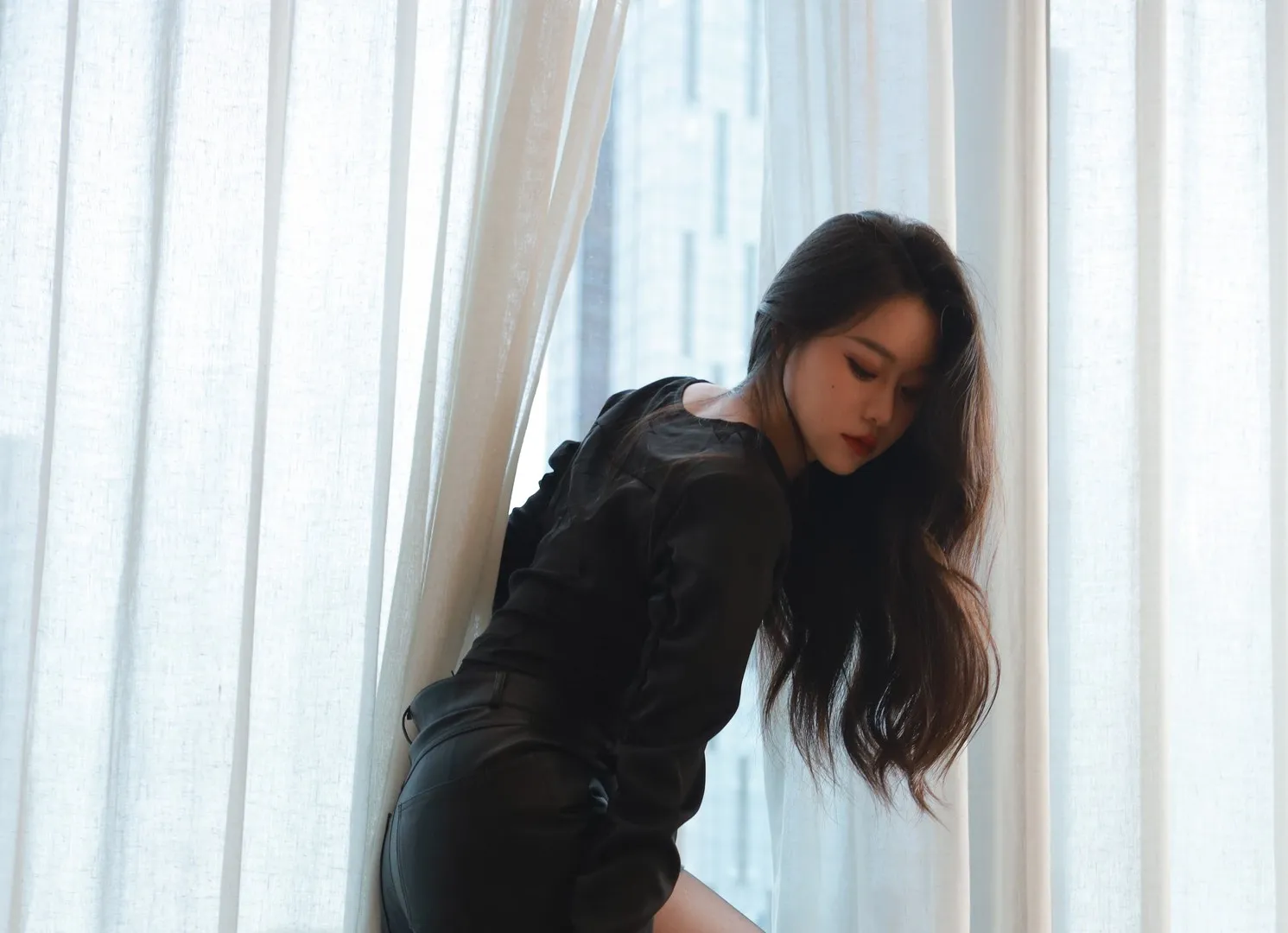 Sia不吃鱼黑色服装写真，人物靠近窗边呈俯身姿态，背景包含白色窗帘和室内空间。